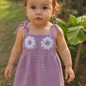 Op de afbeelding: Een lichtpaarse gehaakte jurk met witte bloemenapplicaties. De jurk heeft strikbandjes en een geschulpte zoom. Het kind staat voor een vage groene achtergrond.