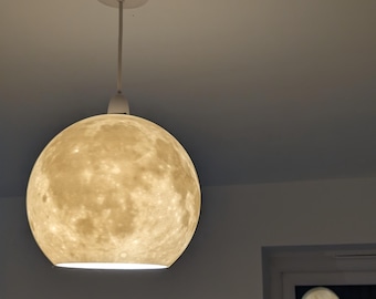 May include: A white ceiling-mounted pendant light with a spherical lampshade. The lampshade is designed to resemble the moon, with a textured, off-white surface. The light fixture is suspended from the ceiling by a white cord.