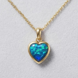 Pode incluir: Um colar de corrente dourada com um pingente em forma de coração que apresenta uma incrustação iridescente em tons vibrantes de azul e verde. O pingente é emoldurado por um engaste dourado liso e suspenso por uma fina corrente de elos.