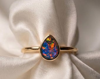 May include: A gold ring featuring a teardrop-shaped opal gemstone. The opal displays a vibrant array of colors, including blue, orange, and green. The ring is set against a backdrop of white fabric, highlighting the jewelry's design.