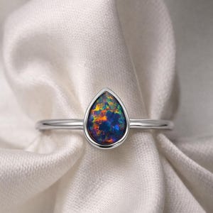 Könnte beinhalten: Ein silberner Ring mit einem tropfenförmigen Opal-Edelstein. Der Opal zeigt eine lebendige Farbpalette, darunter Blau, Orange und Grün. Der Ring ist auf einem weißen, strukturierten Stoff platziert.