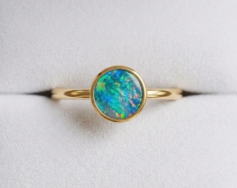 May include: A gold ring featuring a round, bezel-set opal gemstone. The opal displays a vibrant play of color with prominent flashes of blue, green, and orange. The band is thin, polished gold, and the setting holds the stone securely in place.