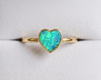 May include: A gold ring featuring a heart-shaped opal stone with vibrant blue and green iridescent flashes. The stone is set in a smooth gold bezel, and the band is a simple, polished gold metal, creating a clean and focused design.