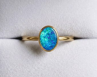 May include: A gold ring featuring an oval-shaped opal gemstone with vibrant blue and green iridescent flashes. The smooth, polished gold band holds the stone in a simple bezel setting, showcasing the play of color within the oval opal center.