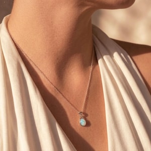 Könnte beinhalten: Eine silberne Halskette mit einer feinen Kette und einem kleinen, ovalen Anhänger, der einen glatten, schillernden blau-weißen Stein enthält. Der Anhänger ist in einer schlichten Metallfassung gefasst und liegt auf dem Schlüsselbein auf.