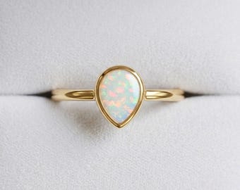 Può includere: Un anello in oro con una pietra di opale a forma di pera incastonata in una lunetta liscia. L'opale mostra un gioco di colori con riflessi rosa, arancioni e verdi su una base bianco latte. La fascia è sottile, lucida e di forma circolare.