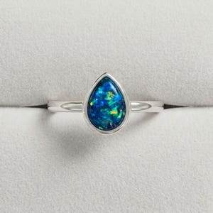 Könnte beinhalten: Ein silberfarbener Ring mit einem birnenförmigen, dunkelblauen Opal-Edelstein mit leuchtend grünen und gelben Sprenkeln. Der Stein ist in einer glatten, polierten Metallfassung gefasst und die Ringschiene besteht aus schlichtem, poliertem Metall.