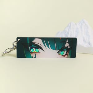 May include: A rectangular keychain featuring a close-up of anime-style eyes. The eyes are a vibrant green with dark teal accents and long lashes. The keychain has a silver-colored clasp.