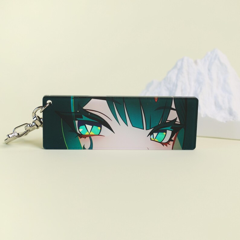 May include: A rectangular keychain featuring a close-up of anime-style eyes. The eyes are a vibrant green with dark teal accents and long lashes. The keychain has a silver-colored clasp.