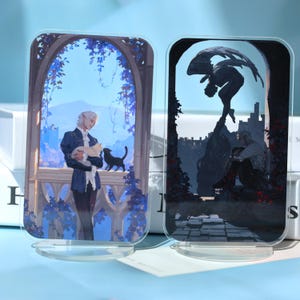 May include: Two acrylic art pieces with illustrations. One features a person with a cat on a balcony, the other depicts a figure with wings. Both are set in a fantasy landscape with blue and black tones.