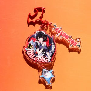 May include: A clear acrylic keychain featuring an anime-style character in a maid outfit, a shark, and the text "Ellen Joe." The keychain has a red bat-shaped top and a small shark charm. The background is orange.