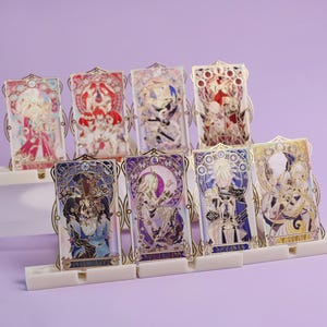 May include: A collection of eight decorative metal art pieces, each featuring a unique character design in vibrant colors. The pieces are displayed in white stands against a soft purple background. The designs have gold-colored frames.