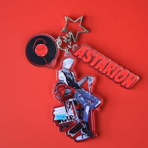 Könnte beinhalten: Ein Schlüsselanhänger mit einer Charakterillustration, einer Vinylplatte und dem Namen "ASTARION" in Rot. Die Figur trägt eine schwarze Weste, zerrissene Jeans und Stiefel und sitzt auf einem Hocker mit dem Text "YOURS BAR". Ein sternförmiger Verschluss verbindet die Anhänger.