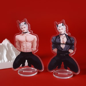 Könnte beinhalten: Zwei Acrylfiguren eines männlichen Charakters mit weißen Haaren und schwarzen Hörnern. Eine Figur ist ohne Hemd, die andere trägt ein schwarzes Hemd. Beide Figuren knien mit den Händen auf dem Rücken. Jede Figur hat einen klaren, runden Sockel.