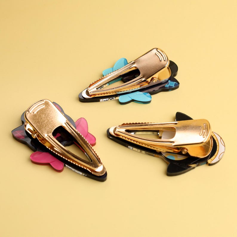 May include: Three gold-colored metal hair clips with decorative bases in pink, blue, and black. The clips are arranged on a yellow background, showcasing their design and color variations. These hair accessories are suitable for various hairstyles.