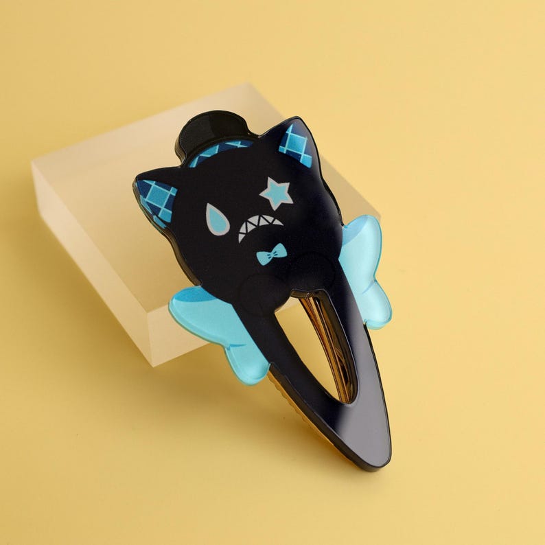 May include: A black hair clip in the shape of a cat with a top hat, star, and bow tie design. The cat has blue accents on its ears and a light blue bow on each side. The clip is made of shiny black plastic.