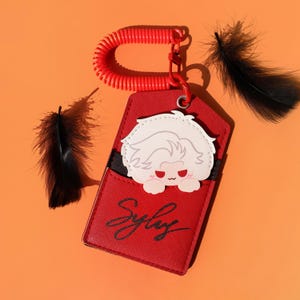 May include: A red keychain with a cartoon character peeking out of a pocket. The keychain has a red coil and a metal clasp. The pocket has the word "Sylby" in a cursive font. Black feathers are scattered around the keychain.
