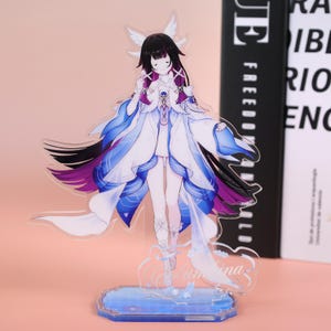 May include: A clear acrylic figurine of a character with black hair, white wings, and a flowing white and blue dress. The figurine stands on a blue base with the name "Valentina" written on it. The character has a serene expression.