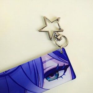 May include: A keychain featuring a silver star-shaped clasp and a rectangular charm. The charm displays a close-up of a character's eye with blue and purple hair and teal eyes. The keychain is on a white background.