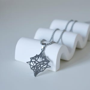 May include: A silver-colored pendant necklace with a geometric design. The pendant is a star-like shape with intricate cutouts, suspended from a delicate chain. The necklace is displayed on a white, wave-shaped stand.