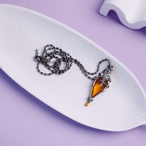Può includere: Una collana con una catena scura e un ciondolo a forma di goccia color ambra. Il ciondolo presenta decorazioni in metallo e una piccola goccia ambrata. La collana è appoggiata su un vassoio bianco a forma di foglia su sfondo viola.