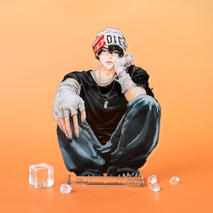 May include: An acrylic figure of a person in a black t-shirt, blue jeans, and a white beanie with the word "DIE" on it. The figure is sitting with their legs crossed, and wearing glasses and fingerless gloves. The background is orange.