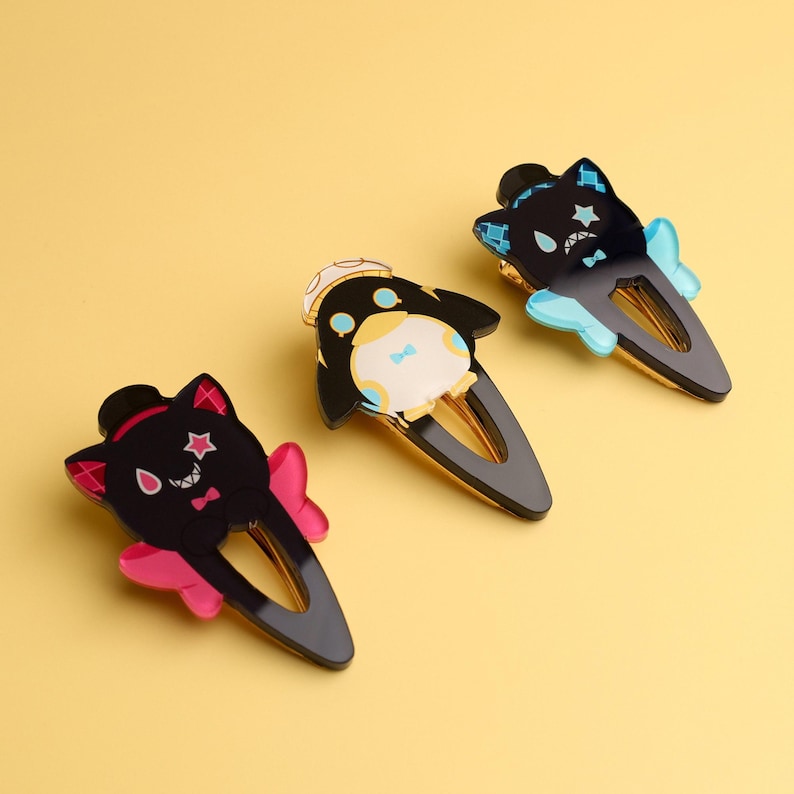 May include: Three decorative hair clips with cartoon cat and penguin designs. The clips feature black, pink, and blue accents, with gold-colored metal clasps. The background is a solid yellow.