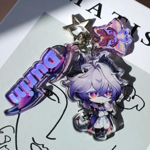 May include: A clear acrylic keychain featuring a cartoon character with white and purple hair, red eyes, and a black and white outfit. The keychain also includes a charm with the word "Durin" in purple and blue, a star-shaped charm, and a floral charm.