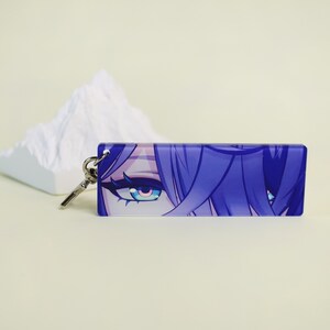 May include: A rectangular keychain featuring a close-up of anime-style eyes with blue and purple hues. The keychain has a silver key ring attached. The background is a light beige color.