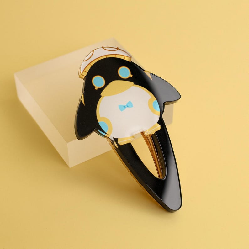 May include: A black and gold penguin-shaped hair clip with a white belly, blue bow tie, and a hat. The clip has a gold-colored base and is set against a yellow background. The penguin has round blue eyes and a yellow beak.
