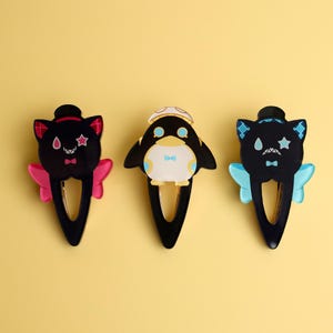 May include: Three decorative hair clips featuring cartoon characters. The clips include a black cat with pink accents, a penguin with a bow tie, and a black cat with blue accents. Each clip has a black base and is set against a yellow background.
