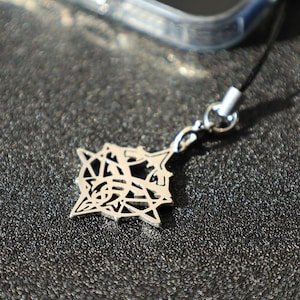 Puede incluir: Un dije plateado con un diseño geométrico, sujeto a un cordón negro. El dije tiene forma de estrella con intrincados patrones recortados. El fondo es una superficie texturizada de color gris oscuro.