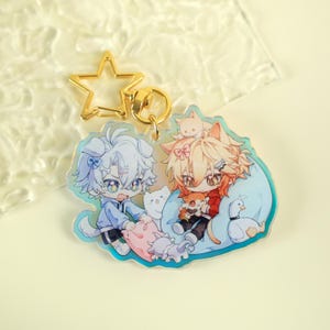 以下が含まれることがあります： 動物の特徴を持つ2人のアニメ風キャラクターが描かれた、透明なアクリルキーホルダー。片方は犬、もう片方は猫です。キーホルダーには金色の星型の留め金と金色のリングが付いています。キャラクターは小さな動物のフィギュアに囲まれています。