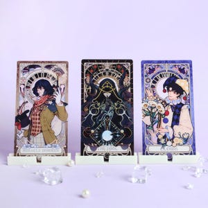 May include: Three tarot card illustrations displayed in white stands. The cards feature detailed artwork with figures and text. The first card reads "MIRACLE INVOKER", the second "THE FOOL", and the third "CLOWN". The cards are set against a light purple background.