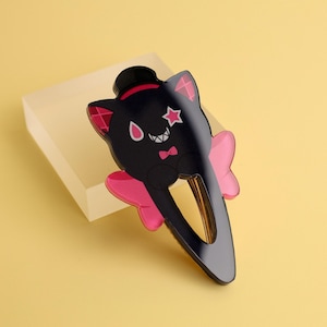 May include: Black cat-shaped hair clip with a top hat, star, and bow tie design. The clip features pink accents on the ears and a bow. The hair accessory is set against a yellow background.
