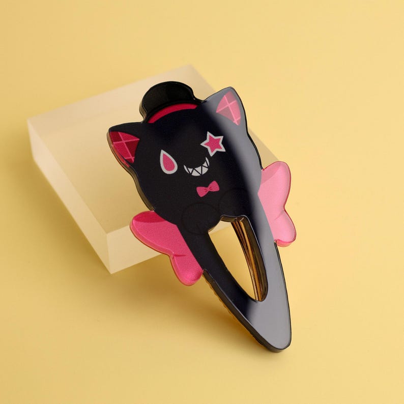 May include: Black cat-shaped hair clip with a top hat, star, and bow tie design. The clip features pink accents on the ears and a bow. The hair accessory is set against a yellow background.