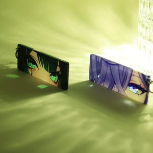 May include: Two rectangular keychains featuring anime-style eye designs. One keychain displays green eyes with black hair, while the other showcases blue eyes with purple hair. Both have a silver keyring attached.