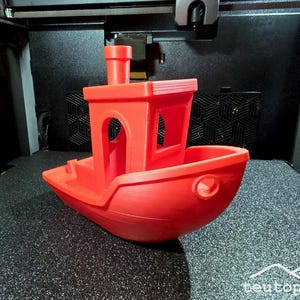 Könnte beinhalten: Ein leuchtend rotes 3D-gedrucktes Spielzeugboot mit Kabine und Schornstein. Das Boot steht auf einer strukturierten schwarzen Oberfläche. Das Bild ist gut beleuchtet und zeigt die Details des Modells.