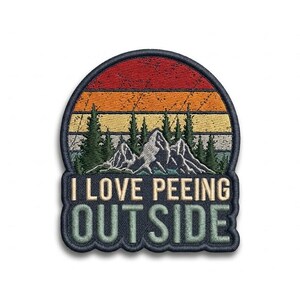May include: An embroidered patch featuring a mountain and forest landscape under a sunset sky with horizontal stripes of red, orange, and yellow. The text I LOVE PEEING OUTSIDE is stitched in white and light green lettering at the bottom.