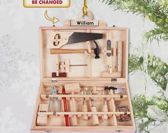 Könnte beinhalten: Ein Werkzeugkasten-Ornament aus Holz mit einer Säge, einem Hammer, einer Zange und verschiedenen Handwerkzeugen in einer rechteckigen Box. Der Deckel ist mit dem Namen William personalisiert. Ein Schild darüber zeigt NAME CAN BE CHANGED.
