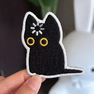 May include: Black cat patch with white border. The cat has yellow eyes and a white starburst design on its head. The patch is held in a hand, with a white rabbit figurine in the background.