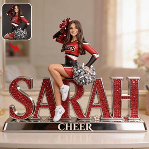 Può includere: Una statuetta personalizzata di cheerleader in rosso, nero e bianco, con il nome "SARAH" in grandi lettere rosse scintillanti. La base recita "CHEER". La cheerleader tiene in mano dei pompon argentati.