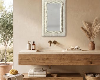 May include: A rectangular wall mirror featuring a thick, cream-coloured, textured fabric frame with a braided border detail. The mirror is mounted on a wall above a wooden vanity with a stone countertop, brass faucet fixtures, and a basin sink.