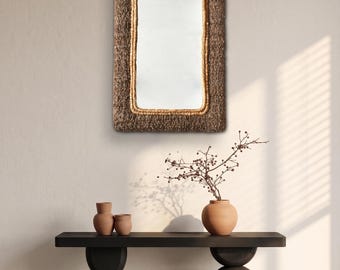 May include: A rectangular wall mirror featuring a thick, textured dark brown woven frame with a lighter tan rope-like border surrounding the reflective glass surface. The mirror is mounted vertically, showcasing a distinct contrast between the materials.