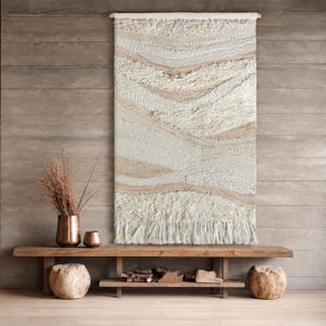 Puede incluir: Un tapiz rectangular para colgar en la pared con un diseño tejido y texturizado en tonos crema, blanco roto y beige. La pieza muestra bandas horizontales onduladas de diversas densidades y acabados, terminando en un largo fleco anudado.