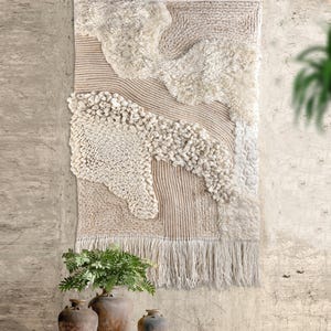 Könnte beinhalten: Ein rechteckiger Wandbehang mit einer Mischung aus cremefarbenen und beigen Texturen. Das Design umfasst Abschnitte aus dickem, zotteligem Garn, eng gewickelten Schlaufen und linearen Rippenmustern. Die Unterkante ist mit langen, losen Fransen versehen.