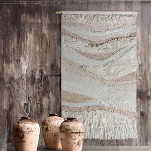 Op de afbeelding: Een geweven wandkleed met een getextureerd, gelaagd ontwerp in crème, beige en lichtbruin. Het rechthoekige stuk heeft horizontale banden en een gefranjerde onderkant. Drie rustieke, lichtbruine vazen met donkere randen staan op de voorgrond.