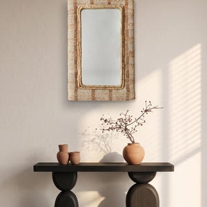 May include: A rectangular wall mirror featuring a thick, textured frame woven from natural fibres in alternating shades of beige and light brown. The mirror has rounded corners and a reflective glass surface centred within the woven border.