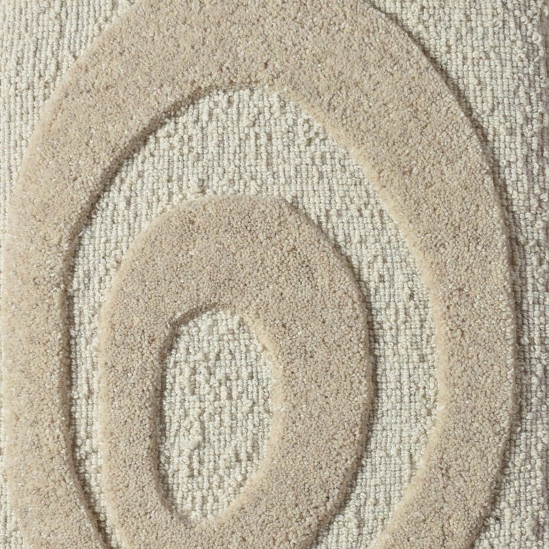 May include: Close-up of a beige rug with a textured surface. The rug features a raised, abstract design of concentric ovals in a lighter beige color. The design adds a modern touch to the neutral-toned rug.