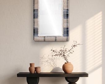 May include: A rectangular wall mirror featuring a thick, woven frame with a plaid pattern in shades of beige, navy blue, and black. The inner edge of the mirror is finished with a textured, cream-coloured braided trim that borders the glass surface.